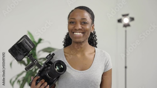 Portrait of Female Videographer or Camera Operator Holding a Video Camera and Smiling Looking Proud. She is on a Video Production Set with a Light and Plant in Background