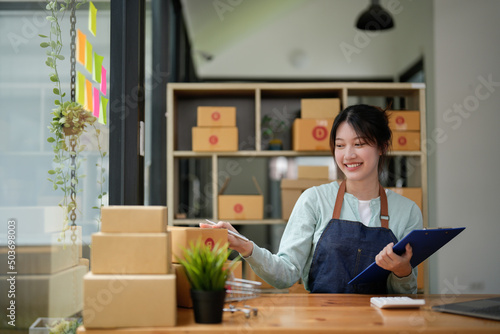 A portrait of Asian woman, e-commerce employee sitting in the office full of packages on the table using a calculator, for SME business, e-commerce, technology and delivery business