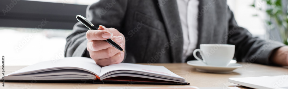 cropped view of banker holding pen near notebook while sitting near coffee cup on blurred background, banner. cropped view of banker holding pen near notebook while sitting near coffee cup on blurred background, banner.