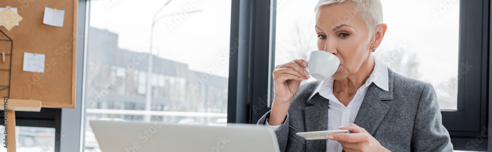 senior economist drinking coffee near blurred laptop in office, banner. senior economist drinking coffee near blurred laptop in office, banner.