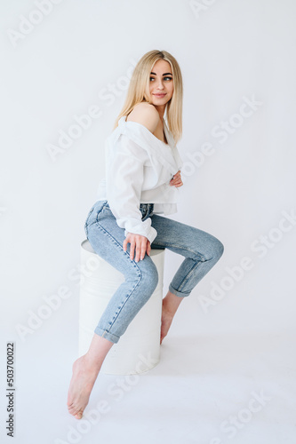A girl in jeans and a white shirt sits on a white barrel. Blonde in jeans on a barrel