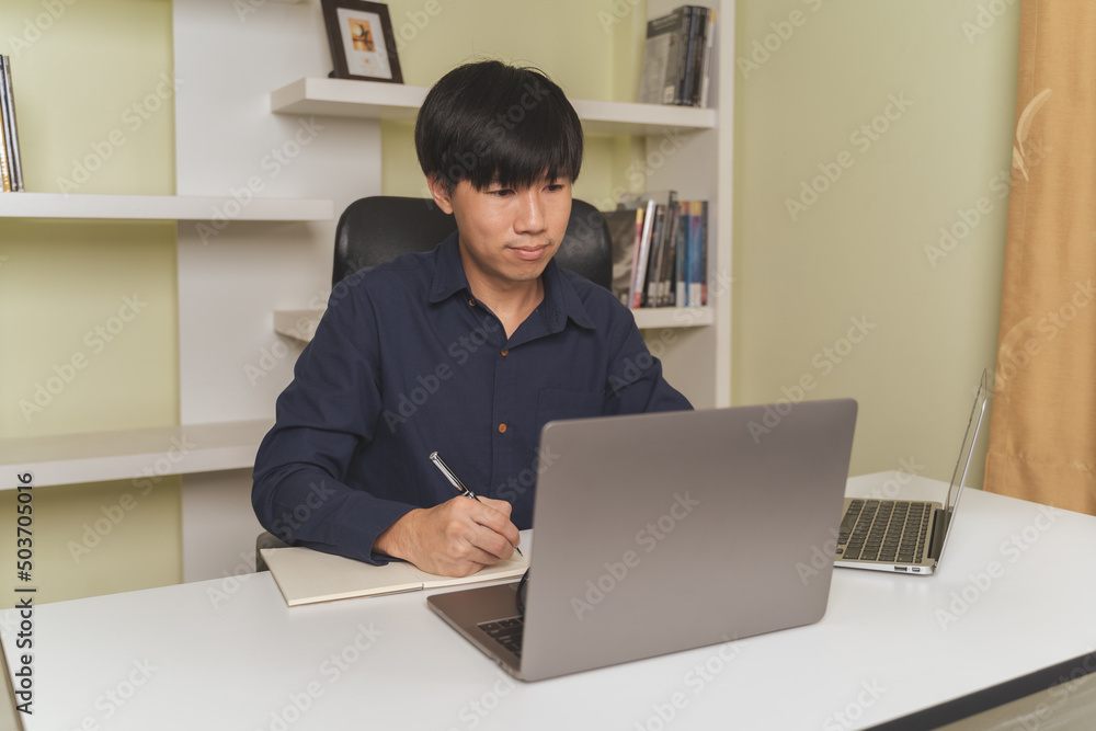 Young Asian college male student writing notes, using laptop for learning online education, business learning, watching online webinar. Businessman is trading, investing on online platform on laptop. Young Asian college male student writing notes, using laptop for learning online education, business learning, watching online webinar. Businessman is trading, investing on online platform on laptop.