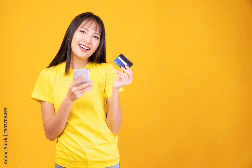 Cheerful smile asian woman holding smart phone and mockup credit card for internet banking while standing over isolated yellow background. Financial payment online shopping and money transfer concept. Cheerful smile asian woman holding smart phone and mockup credit card for internet banking while standing over isolated yellow background. Financial payment online shopping and money transfer concept.