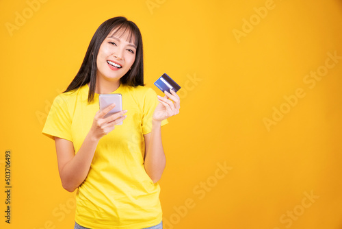 Cheerful smile asian woman holding smart phone and mockup credit card for internet banking while standing over isolated yellow background. Financial payment online shopping and money transfer concept.