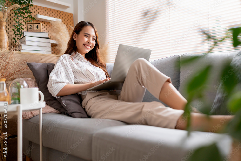 young attractive asian female working at home hand use laptop checking search schedule working process planning report with leisure relax on sofa in living room with morning light peaceful moment - obrazy, fototapety, plakaty 