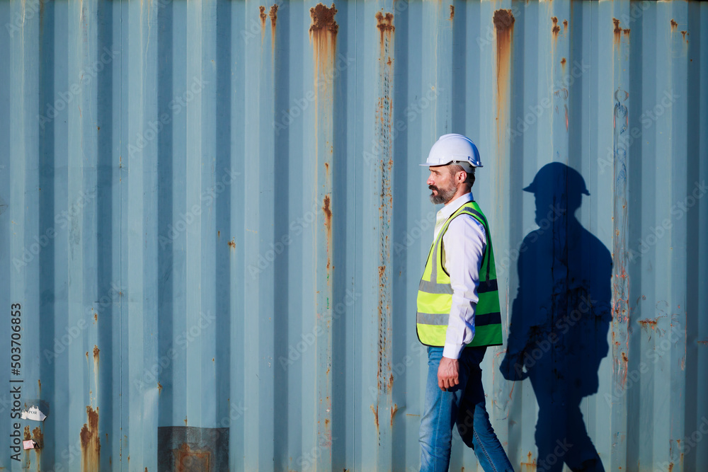 Male walk. Handsome caucasian male dock foreman worker in white hard hat helmet and high-visibility vest walking in Shipping Cargo Container Terminal Depot Male walk. Handsome caucasian male dock foreman worker in white hard hat helmet and high-visibility vest walking in Shipping Cargo Container Terminal Depot