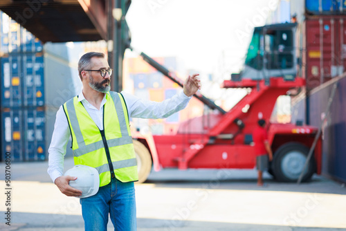 Handsome caucasian male dock foreman worker in white hard hat helmet and high-visibility vest working in Shipping Cargo Container Terminal Depot