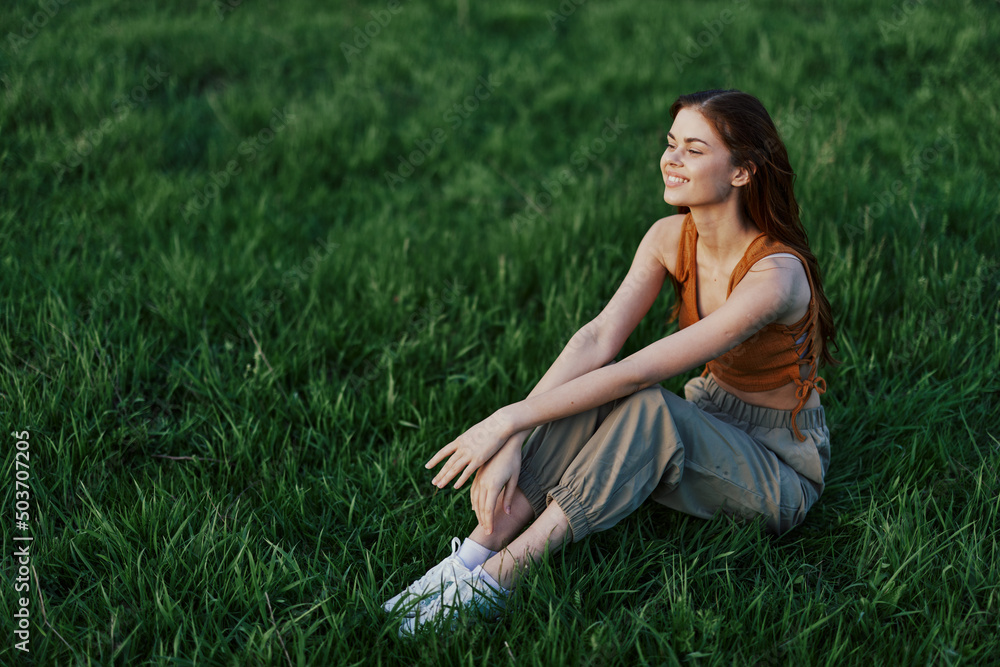 A beautiful girl relaxing in nature sitting in pants and top on the green grass in the rays of the setting summer sun. The concept of health and care for the body and the environment A beautiful girl relaxing in nature sitting in pants and top on the green grass in the rays of the setting summer sun. The concept of health and care for the body and the environment