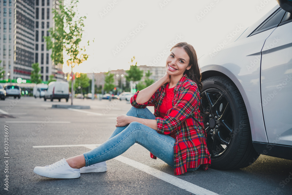 Fototapeta premium Portrait of attractive cheerful girl driver sitting on floor near new modern car ownership dealer on parking outdoors