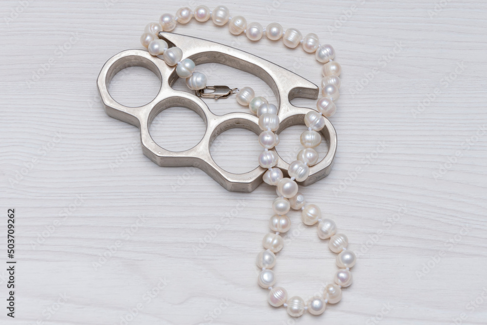 Obraz premium A pearl necklace and a metal brass knuckles lie on a light textured wooden background. Concept: women's accessories, robbery on the street or at home.