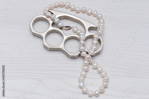 A pearl necklace and a metal brass knuckles lie on a light textured wooden background. Concept: women's accessories, robbery on the street or at home.