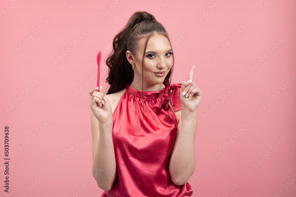 Portrait of cheerful woman with attractive smile, with ponytail of hair in elegant blouse isolated on pink background, holding pen and have great idea. Beautiful woman showing emotion.