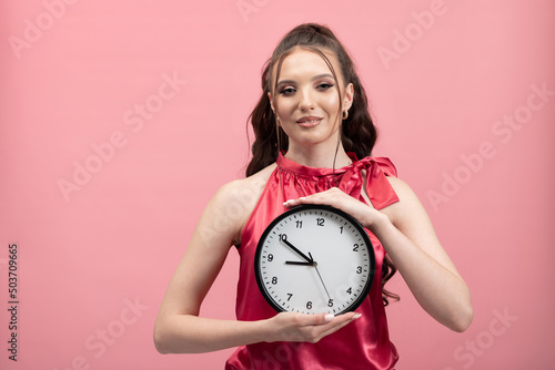 Young smiling woman in pink jacket holds clock portrait on pastel background studio portrait. Concept of time management, punctuality.