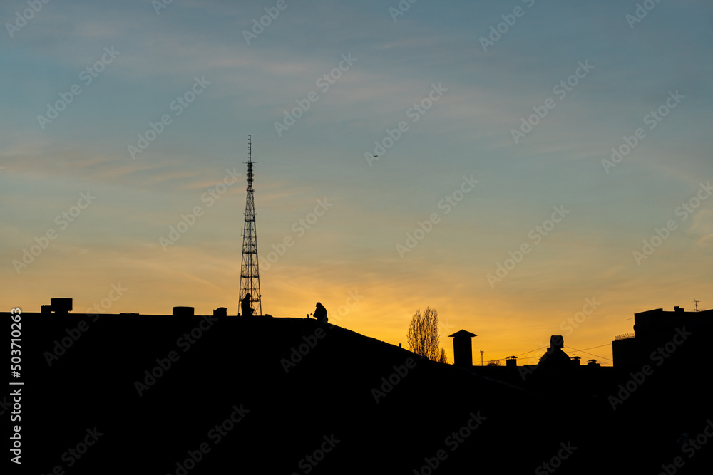 A silhouette of two men working on a roof of a building in the city at a sunset time. Golden hour, big tower and residential buildings.
