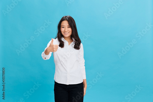 Joyful cute Asian businesswoman in classic office dress code show thumb up gesture Like posing isolated on over blue studio background. Cool business offer. People Emotions Business concept