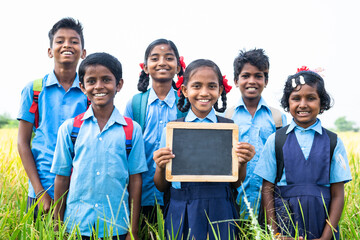 Village teenage kids showing empty slate board by looking at camera at farmland - concept of advertisement, education and friendship