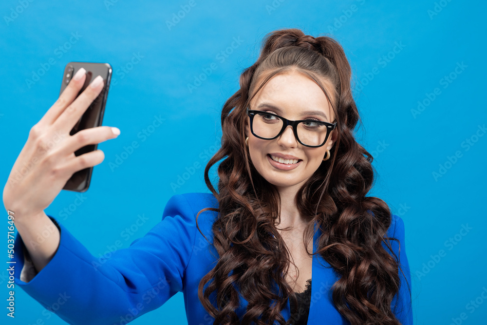 Portrait of a cheerful, positive, smiling woman holding a phone, earphones in hands, taking a selfie against a blue background.