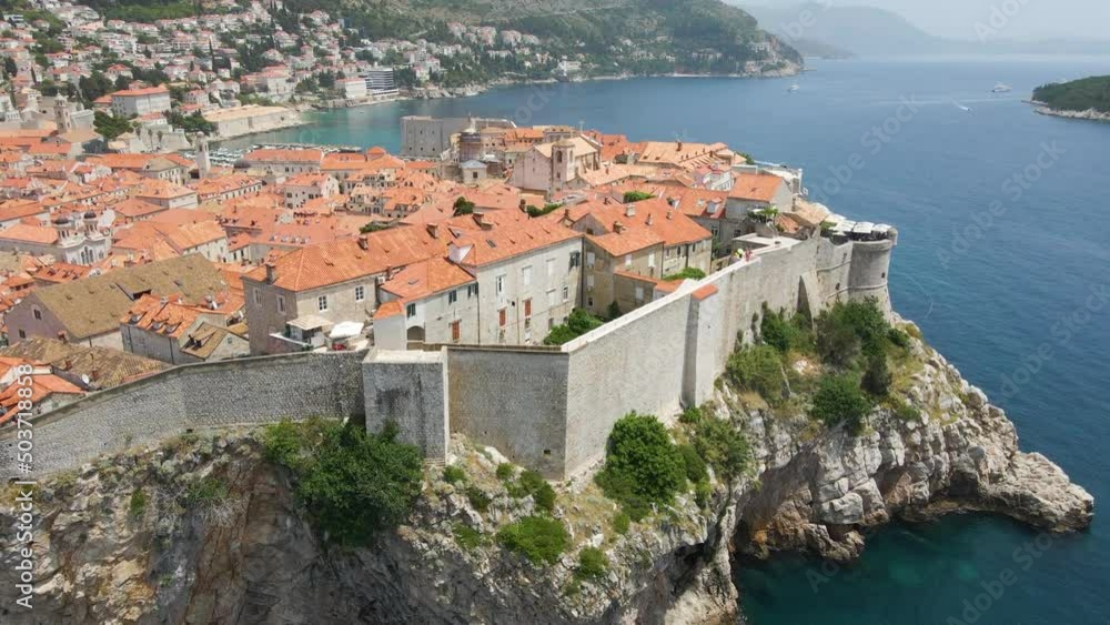 Aerial drone forwarding shot over the old fortress wall along the old town of Dubrovnik, Croatia with the view of building rooftops on a sunny daytime. Rocky cliffs. Aerial drone forwarding shot over the old fortress wall along the old town of Dubrovnik, Croatia with the view of building rooftops on a sunny daytime. Rocky cliffs.