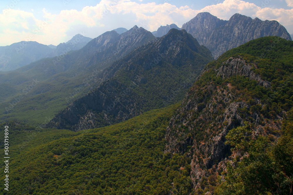 Obraz premium Mountain peaks overgrown with trees and bushes on a background of blue sky with clouds from Mount Tünektepe on the outskirts of Antalya in southern Turkey 