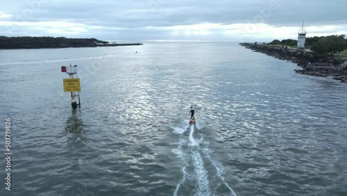 A jet board carves through the glassy morning waters along a city harbor break wall towards a dolphin. Drone view photo