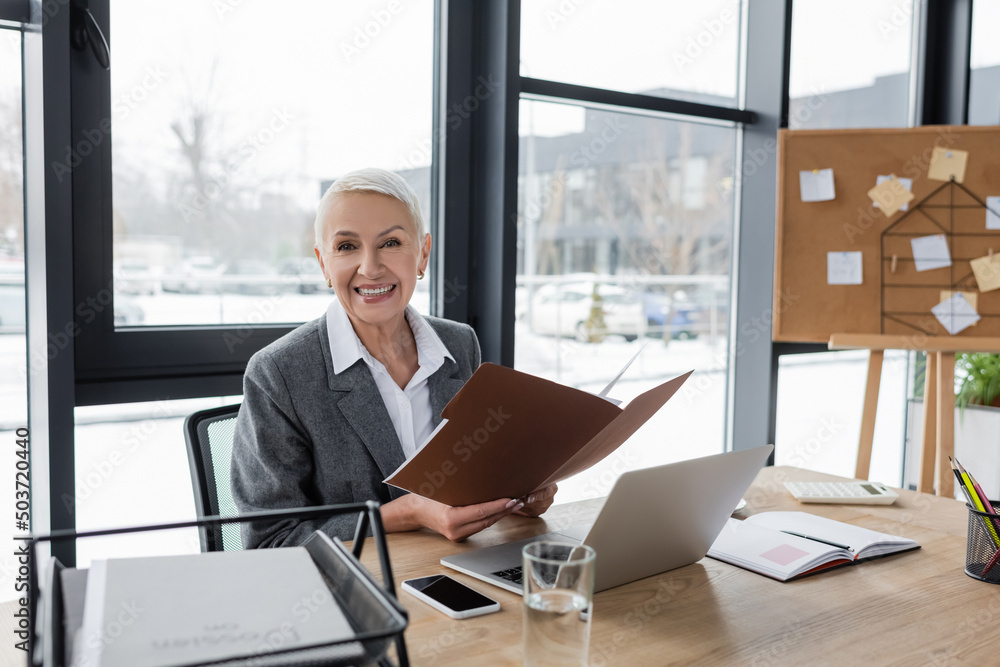happy banker with folder smiling at camera near laptop and cellphone with blank screen. happy banker with folder smiling at camera near laptop and cellphone with blank screen.