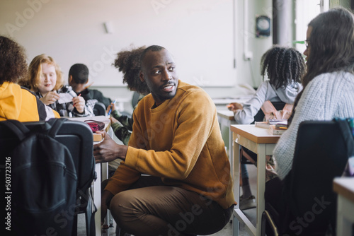 Side view of male teacher talking with student while crouching amidst desks in classroom photo