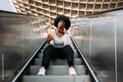 Young black teenage woman with afro hair sitting on an outdoor escalator taking a selfie blowing a kiss.