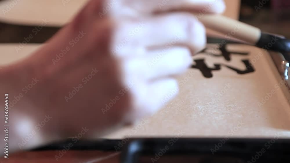 Close-up of a hand and a calligraphy brush. A person learns to hold a brush and draw elements of hieroglyphs on paper. Multiple attempt to draw the hieroglyph - a woman with a child. Close-up of a hand and a calligraphy brush. A person learns to hold a brush and draw elements of hieroglyphs on paper. Multiple attempt to draw the hieroglyph - a woman with a child.