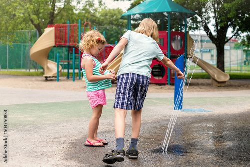 Little happy children playing at water splash pad fountain in park playground on hot summer day. Kids having fun outdoors in summertime.