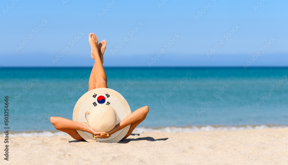 Fototapeta premium A slender girl on the beach in a straw hat in the colors of the South Korea flag. The concept of a perfect vacation in a resort in the South Korea. Focus on the hat.