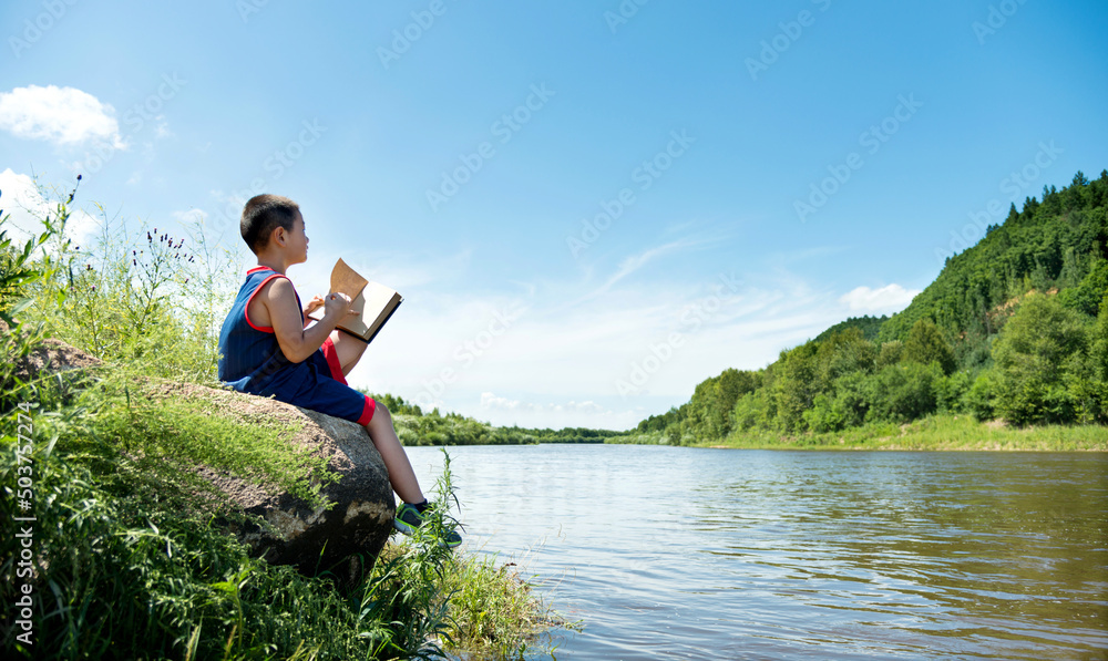 Little boy enjoying reading a book by the lake