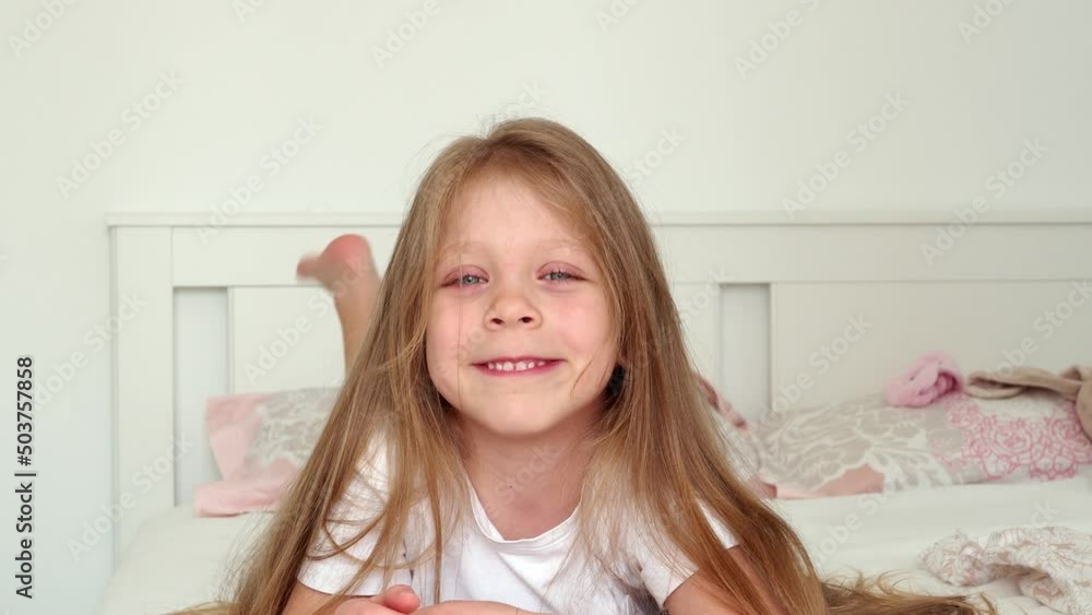 portrait of a smiling little girl. A beautiful, sweet little girl with blue eyes and long hair lies on her stomach in bed and smiles. The child is naughty and indulges. Children's pranks and games in portrait of a smiling little girl. A beautiful, sweet little girl with blue eyes and long hair lies on her stomach in bed and smiles. The child is naughty and indulges. Children's pranks and games in