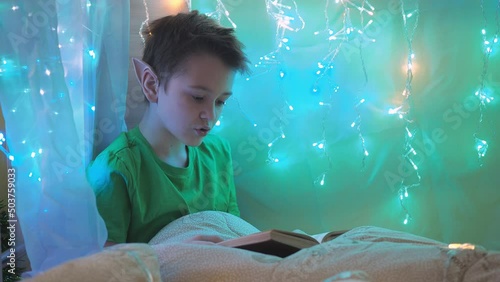 a teenager with long elf ears reads a book sitting on a bed decorated with garlands. wrapped in a blanket