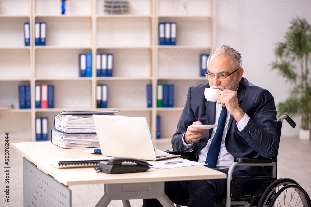 Old male employee in wheel-chair sitting at workplace Old male employee in wheel-chair sitting at workplace