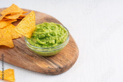 Homemade organic green mexican guacamole dip, sauce or spread made of ripe mashed avocado served in glass bowl with nachos or tortilla chips on cutting board on white wooden background with copy space