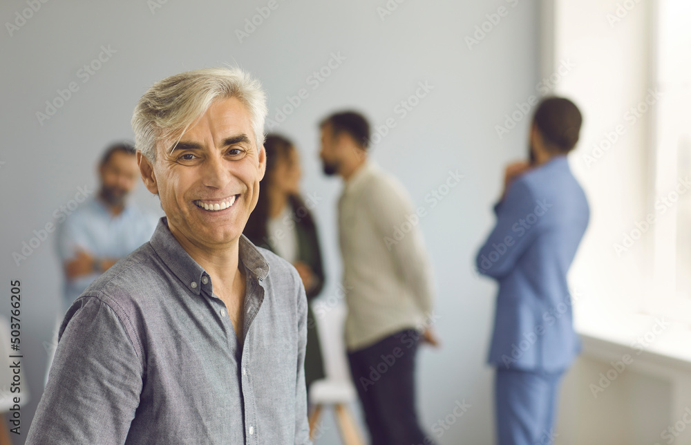 Portrait of happy successful mature business leader and company owner. Positive handsome white haired senior Caucasian man looking at camera and smiling standing against blurred office background Portrait of happy successful mature business leader and company owner. Positive handsome white haired senior Caucasian man looking at camera and smiling standing against blurred office background