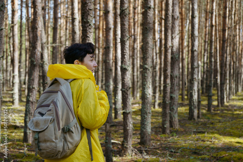 A young woman in a yellow raincoat and with a backpack stands in the woods with her back to the camera