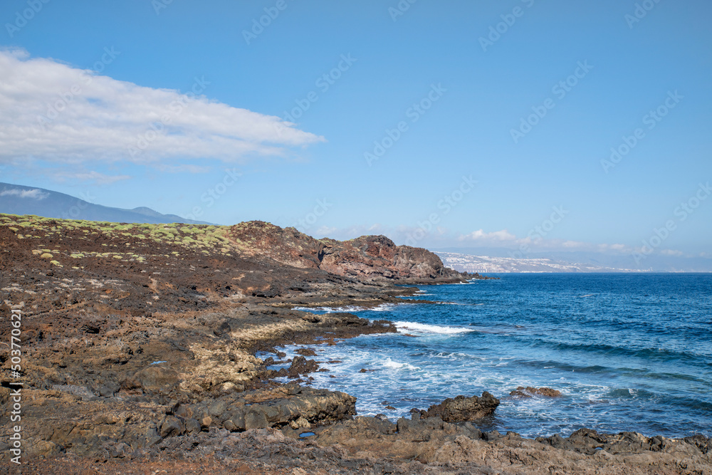Path through the rough volcanic lands known as malpais, at Puertito de Guimar, with clear ocean waves and views of mountain range and El Socorro town in the distance, Tenerife, Canary Islands, Spain Path through the rough volcanic lands known as malpais, at Puertito de Guimar, with clear ocean waves and views of mountain range and El Socorro town in the distance, Tenerife, Canary Islands, Spain