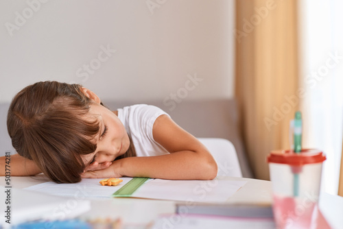 Indoor shot of dark haired female kid wearing white t shirt sleeping at table, being tired while doing homework, laying on desk, being exhausted and overworked.