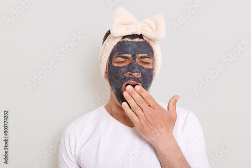 Portrait of funny man wearing white t shirt and female's hair band standing with mud mask and yawning, covering mouth with palm, being tired to wait result posing isolated over gray background.