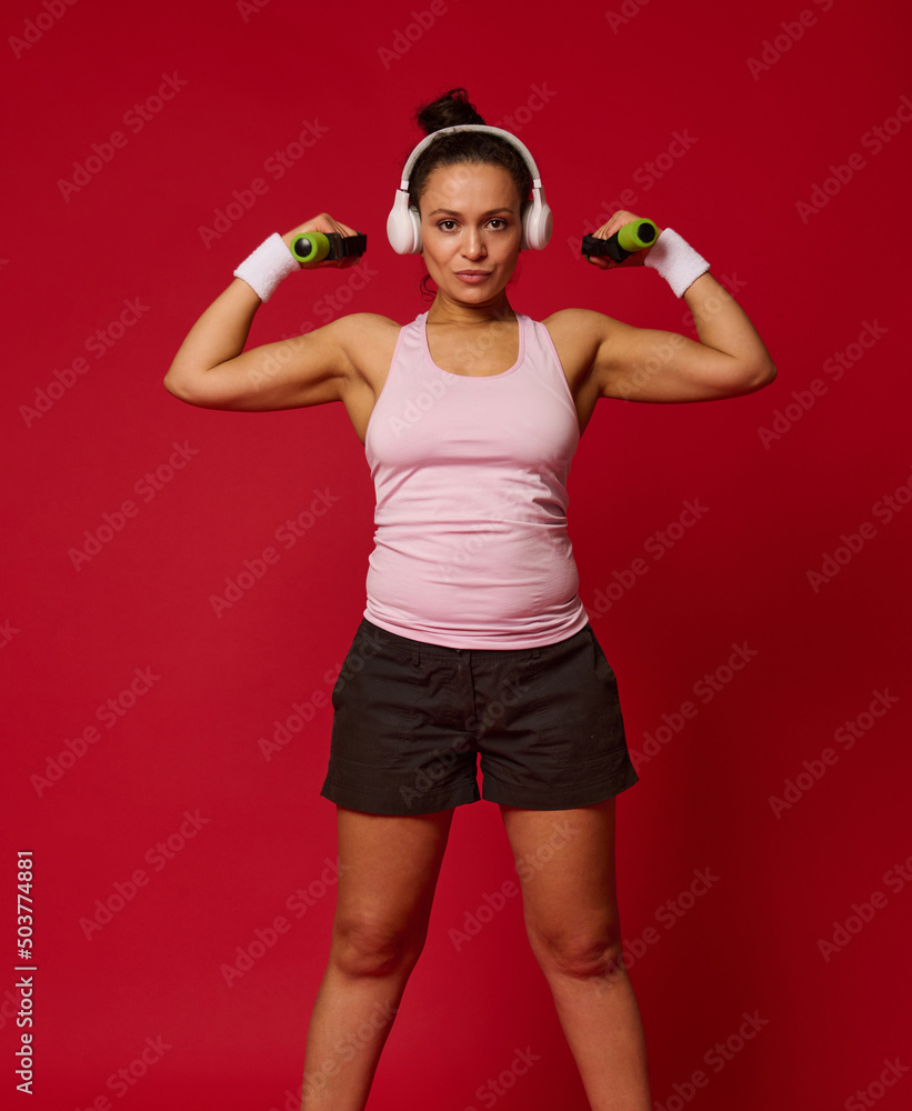 Middle-aged active multi-ethnic woman exercises with dumbbells on red colored background. Sport, fitness, motion, motivation, body positivity concept Middle-aged active multi-ethnic woman exercises with dumbbells on red colored background. Sport, fitness, motion, motivation, body positivity concept
