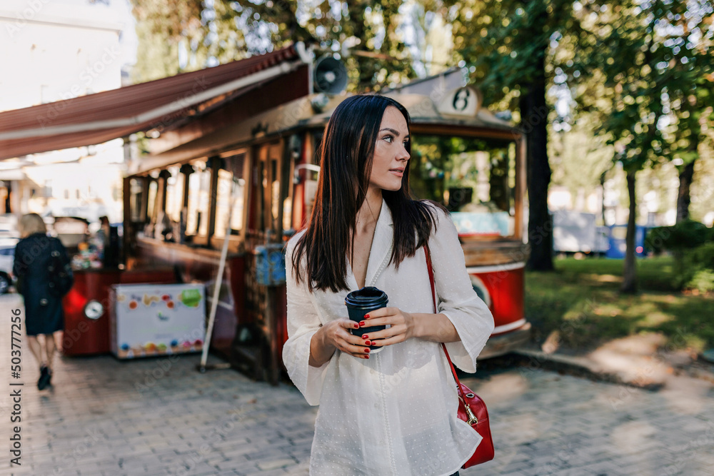 Fototapeta premium Stylish spectacular lady with dark hair wearing white shirt is holding cup with coffee is looking aside on city park background in good mood. 