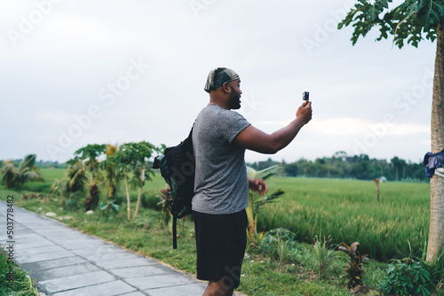 Fototapeta Naklejka Na Ścianę i Meble -  Male videographer with touristic backpack using gopro camera equipment for shooting influence vlog about travel getaway journey in Indonesia, man with waterproof technology talking about trip