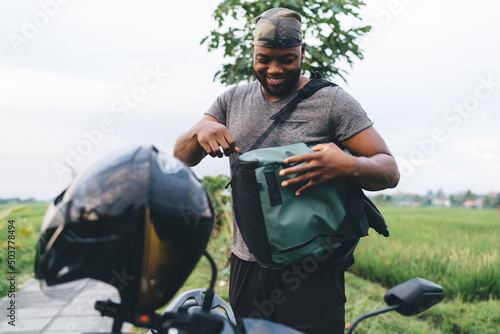 Cheerful African American tourist smiling near scooter with helmet while searching something in backpack at Bali, happy dark skinned hipster guy in bandanna enjoying summer vacations in Indonesia #503778494