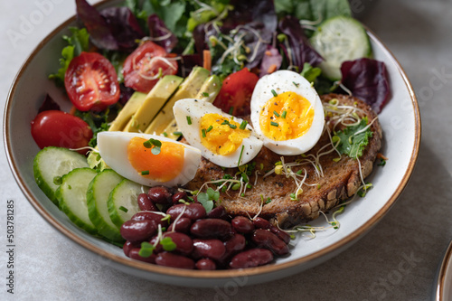 Light green salad with leaves, avocado, cherry tomatoes, cucumber slices, red beans, micro greens and medium boiled eggs on slice of toasted bread.
