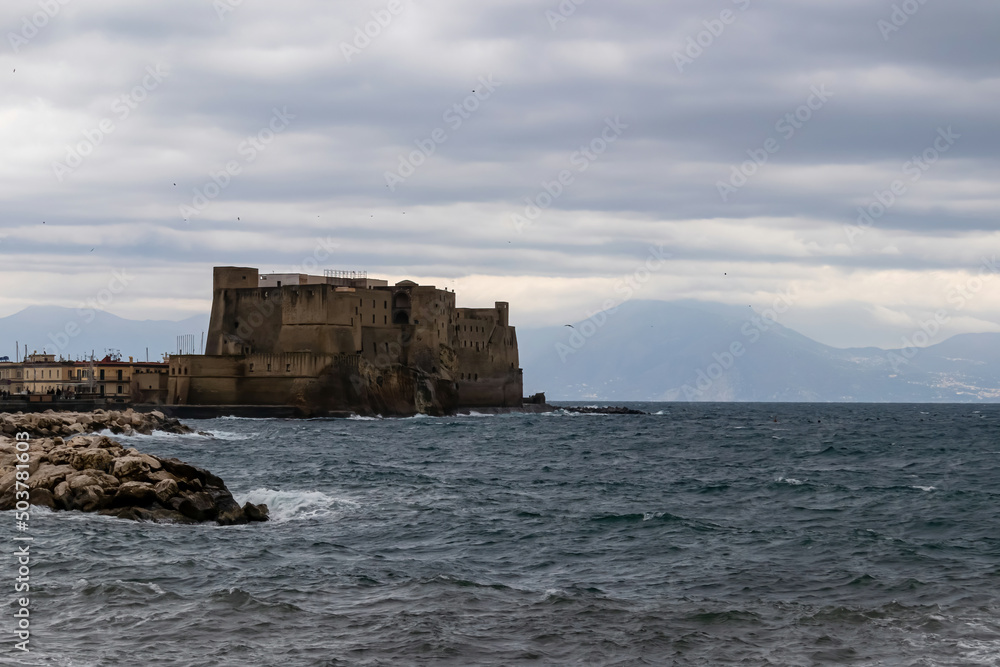 Panoramic view on Castel dell Ovo (Egg Castle) on a cloudy winter day in Naples, Campania, Italy, Europe. Clouds are accumulating around nearby mountains. Sea is calm. Walking along the promenade