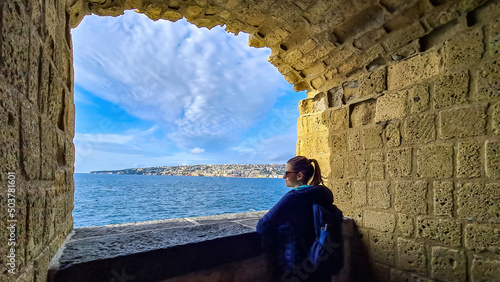 A tourist woman enjoying the panoramic view from Castel dell Ovo (Egg Castle) on the city of Naples, Campania, Italy, Europe. Person leaning against the castle wall, bathing in the sun. Sea view photo