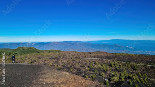 Tourist man hiking around the crater of Sartorio on volcanic landscape of volcano mount Etna, in Sicily, Italy, Europe. View on the Ionian sea. Walking on solidified lava, ash and pumice ground