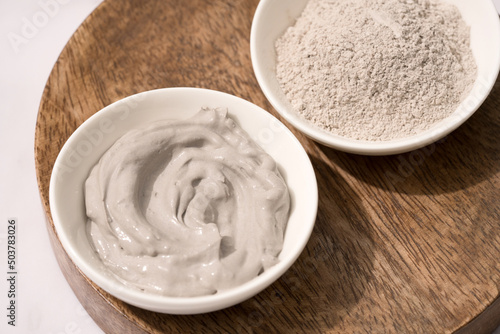 Bowl with gray cosmetic clay cream and clay powder on wooden tray - mineral bentonite facial mask. Skincare beauty concept. Selective focus