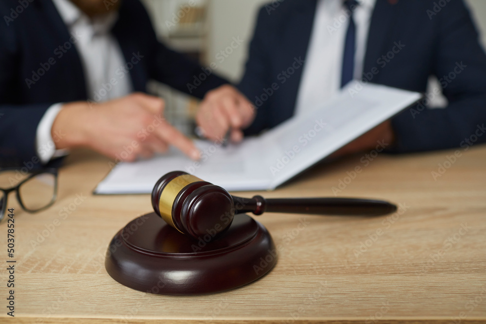 Judge's gavel on wooden desk up close, and two people discussing something in background. Lawyer or attorney giving consultation and helping client with documents. Law services, legal advice concept Judge's gavel on wooden desk up close, and two people discussing something in background. Lawyer or attorney giving consultation and helping client with documents. Law services, legal advice concept